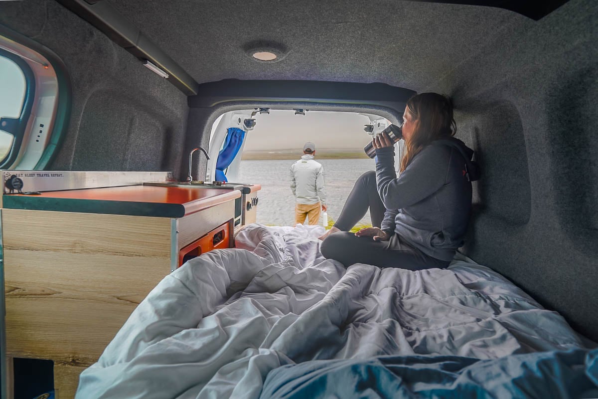 Woman sipping a coffee thermos while looking out the backdoor of a campervan with a man in the background looking over the ocean in Iceland