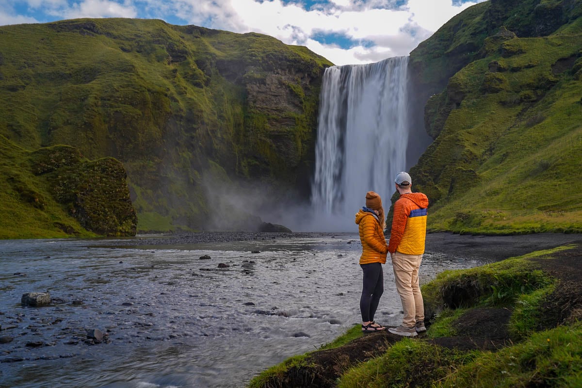 Couple standing at the base of Skogafoss with mossy mountains in the background in Iceland