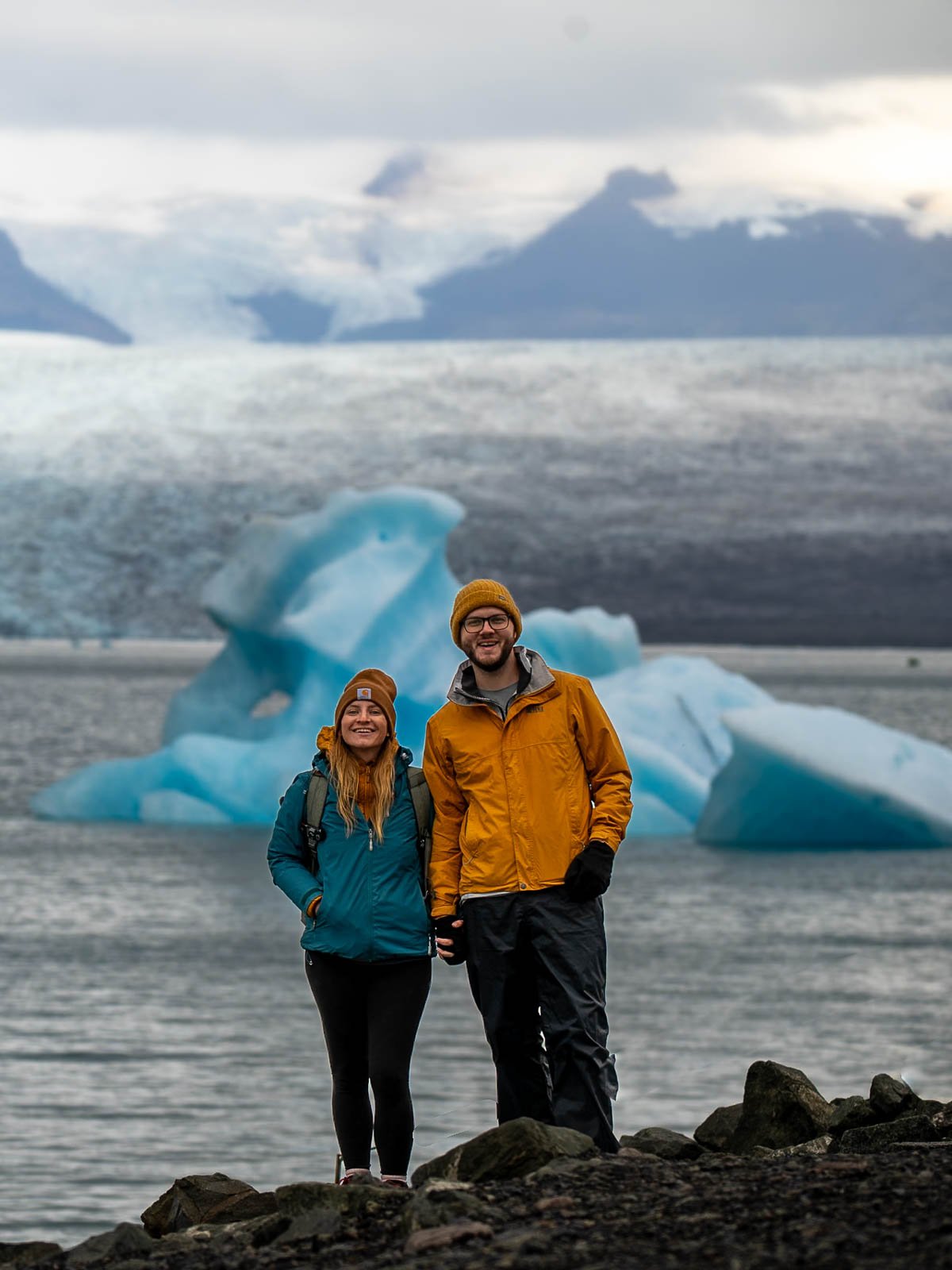 Couple smiling in front of a blue iceberg with a glacier in the background at the Jökulsárlón Glacier Lagoon in Iceland