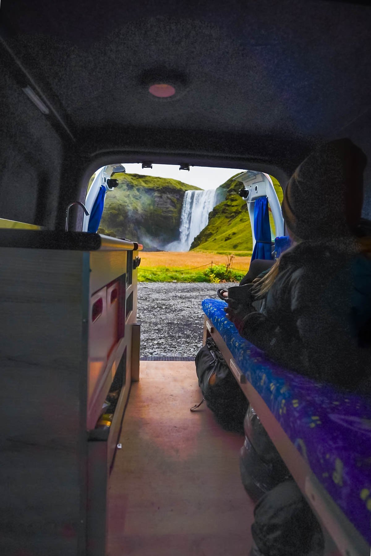 Woman laying in the back of a campervan with open doors with Skogafoss in the background in Iceland