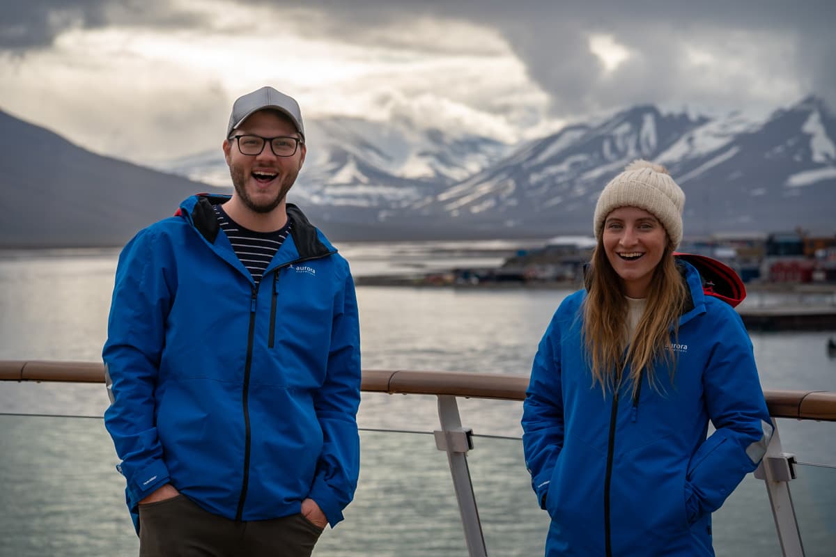 Couple smiling on the top deck of a cruise ship with colorful buildings of Longyearbyen, Svalbard and snowy mountains in the background