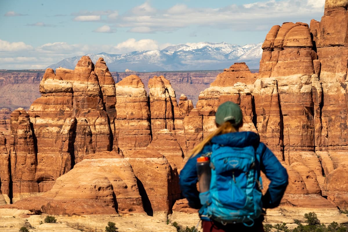 Woman with a backpack looking at striated rock formations with a mountain in the distance along the Druid Arch Trail in Canyonlands National Park in Utah