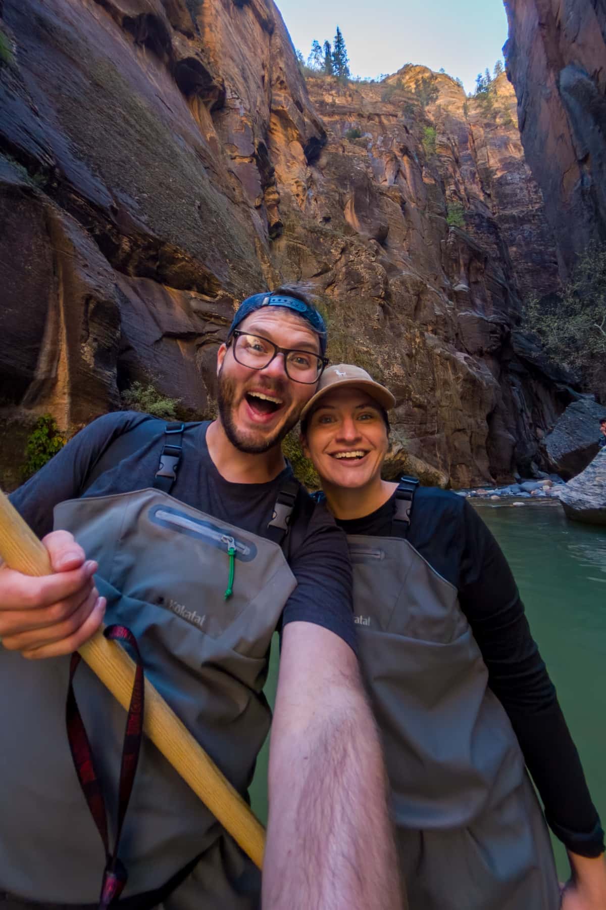 Couple smiling while wearing waders and holding hiking poles in The Narrows in Zion National Park