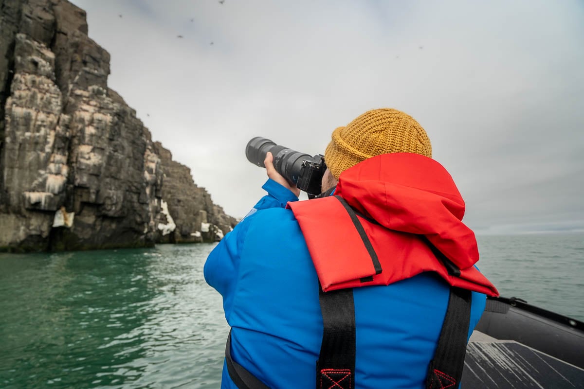 Man on a Zodiac boat with a long camera lens with volcanic cliffside in the background in Svalbard, Norway