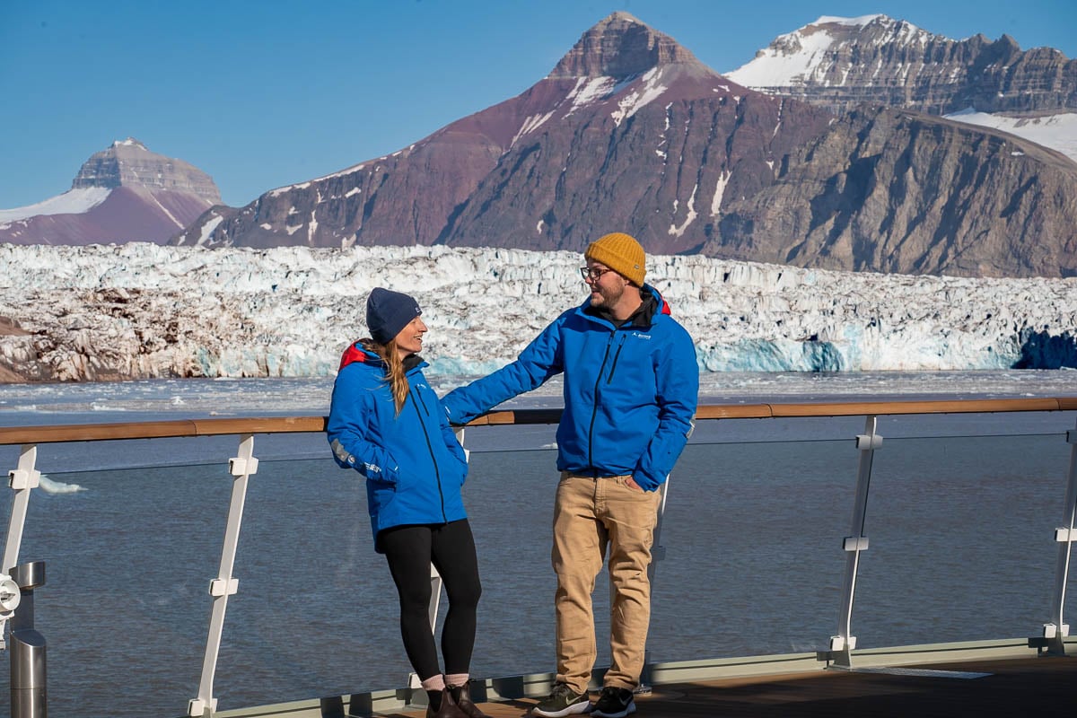 Couple leaning against the railing on the Observation Deck with a glacier and mountains in the background in Svalbard, Norway