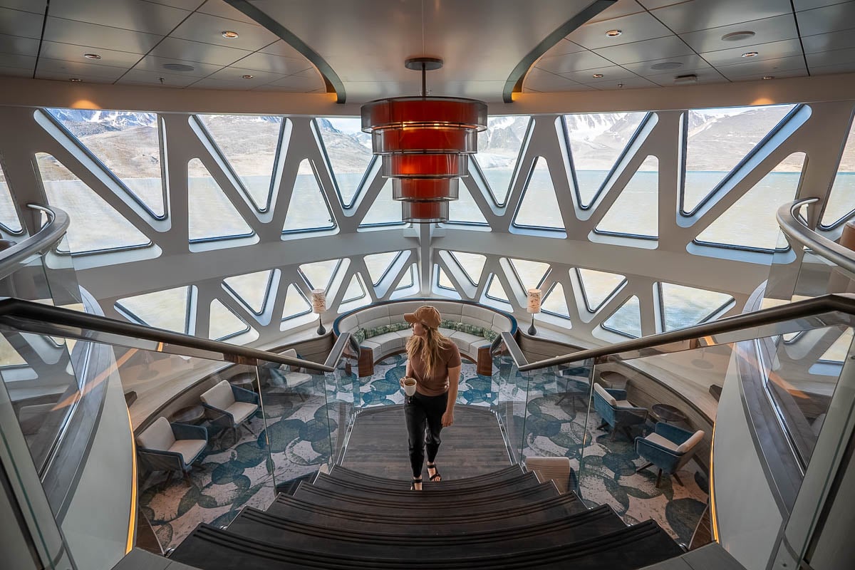 Woman holding a mug walking up stairs in the library of the Sylvia Earle cruise ship with mountains out the window in Svalbard, Norway