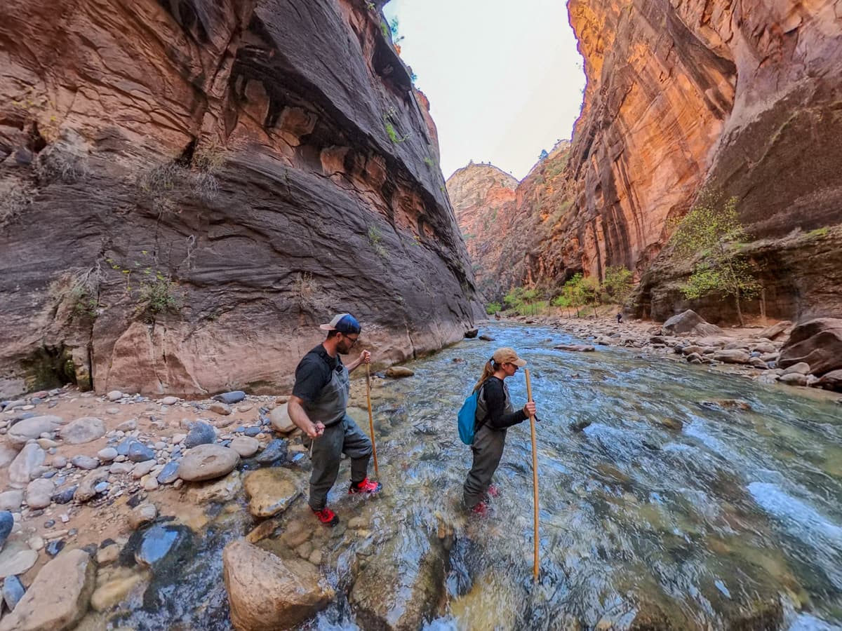 Couple walking across the Virgin River with walls of Zion Canyon in the background along The Narrows in Zion National Park