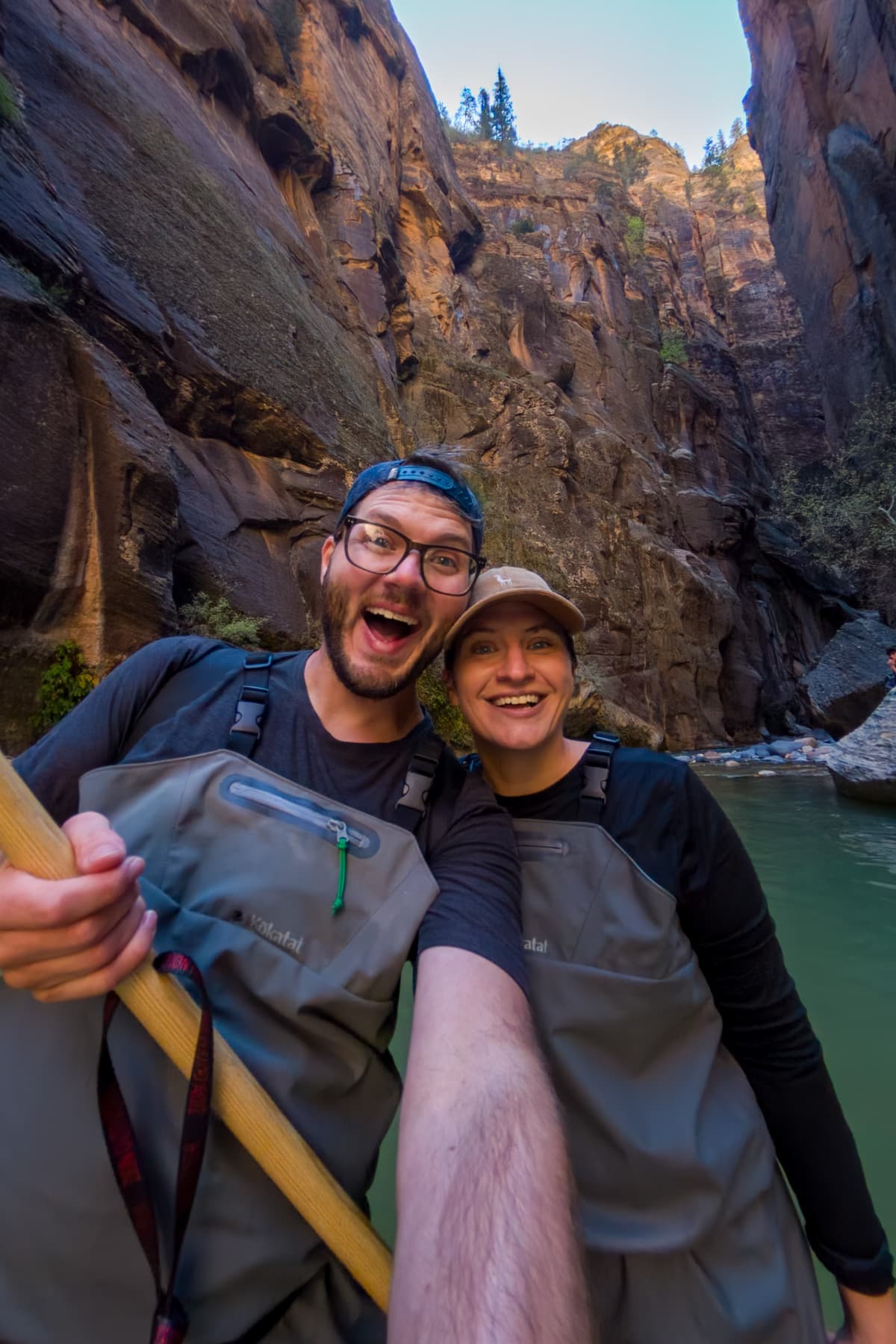 Smiling couple wearing waders standing in the Virgin River with canyon walls in the background along The Narrows trail in Zion National Park