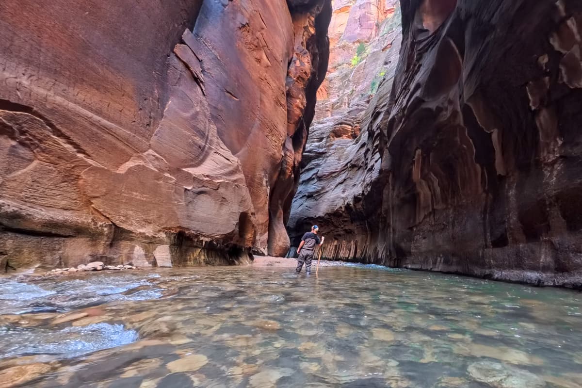 Man standing in waders looking up at walls of the Zion Canyon while hiking along The Narrows in Zion National Park