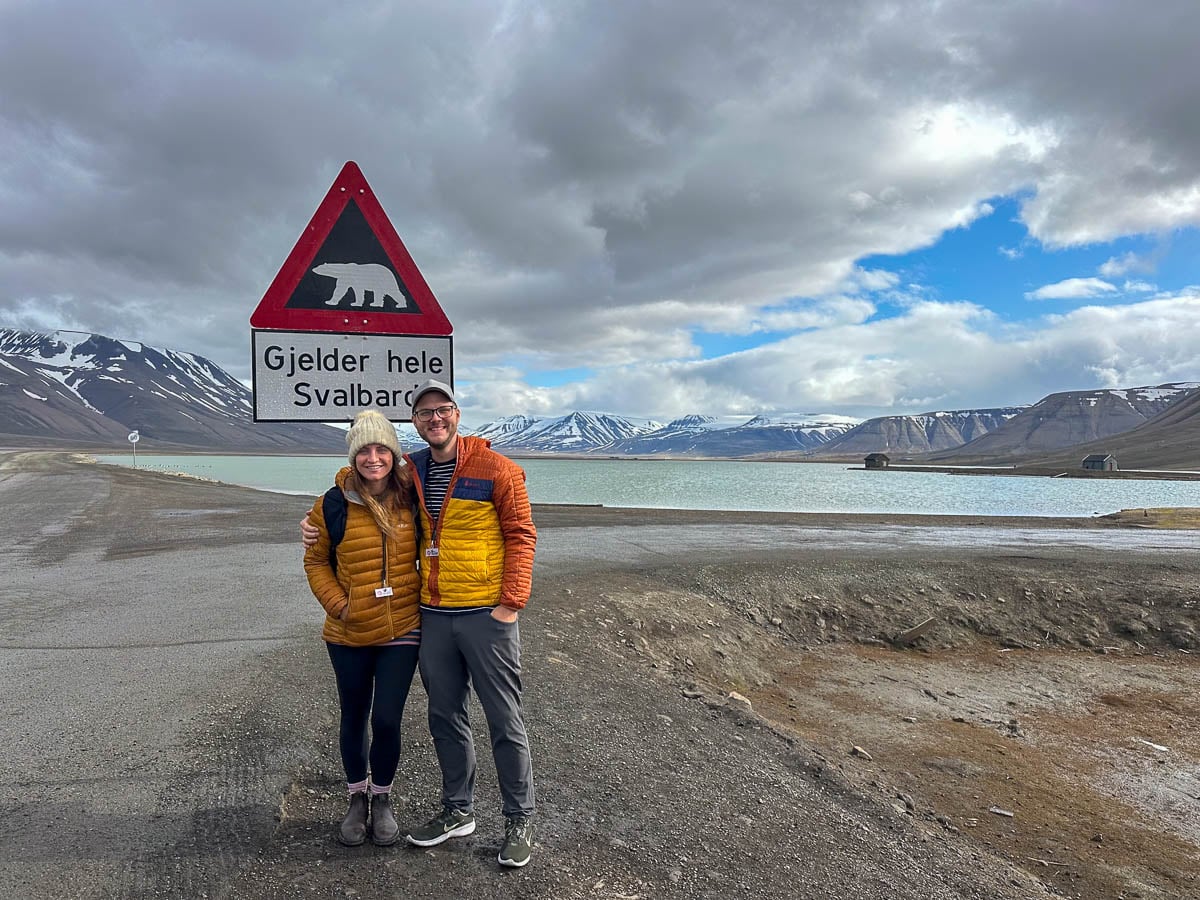 Couple smiling in front of a polar bear warning sign in Svalbard, Norway with fjords and mountains in the background