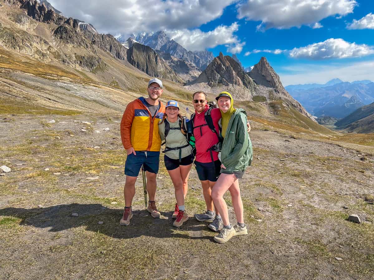 Four smiling friends at the Col de la Seigne pass with jagged mountains in the background along the Tour du Mont Blanc