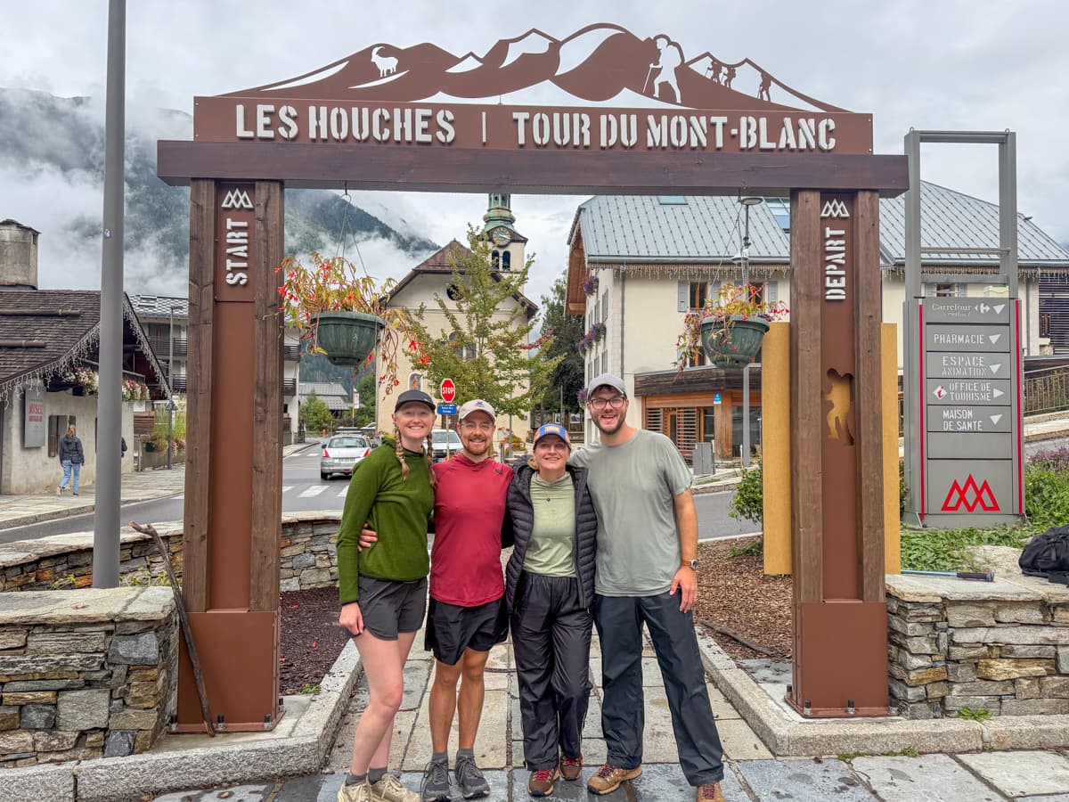 Four smiling friends at the Tour du Mont Blanc starting arch in Les Houches, France