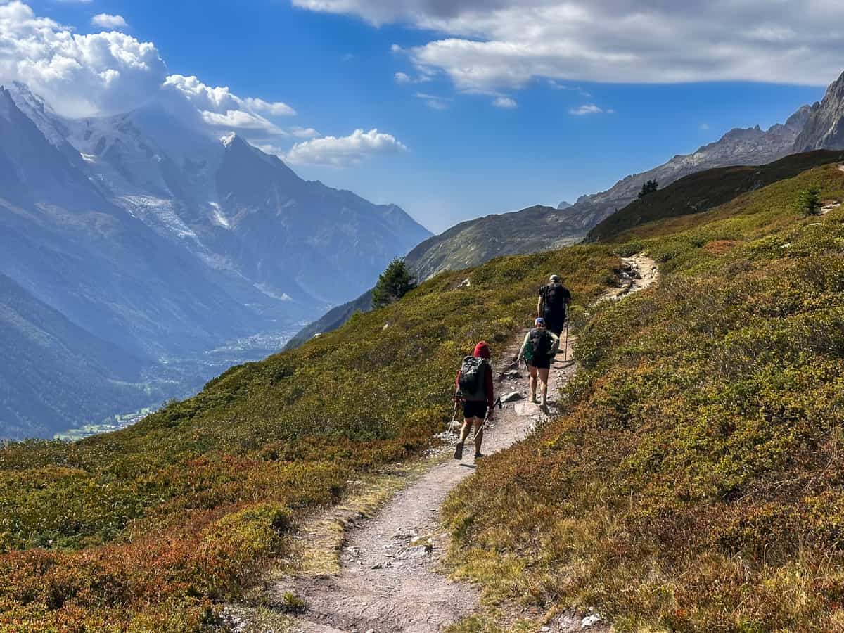 Three hikers hiking through a grassy meadow with the Mont Blanc massif in the background outside of Chamonix, France along the Tour du Mont Blanc