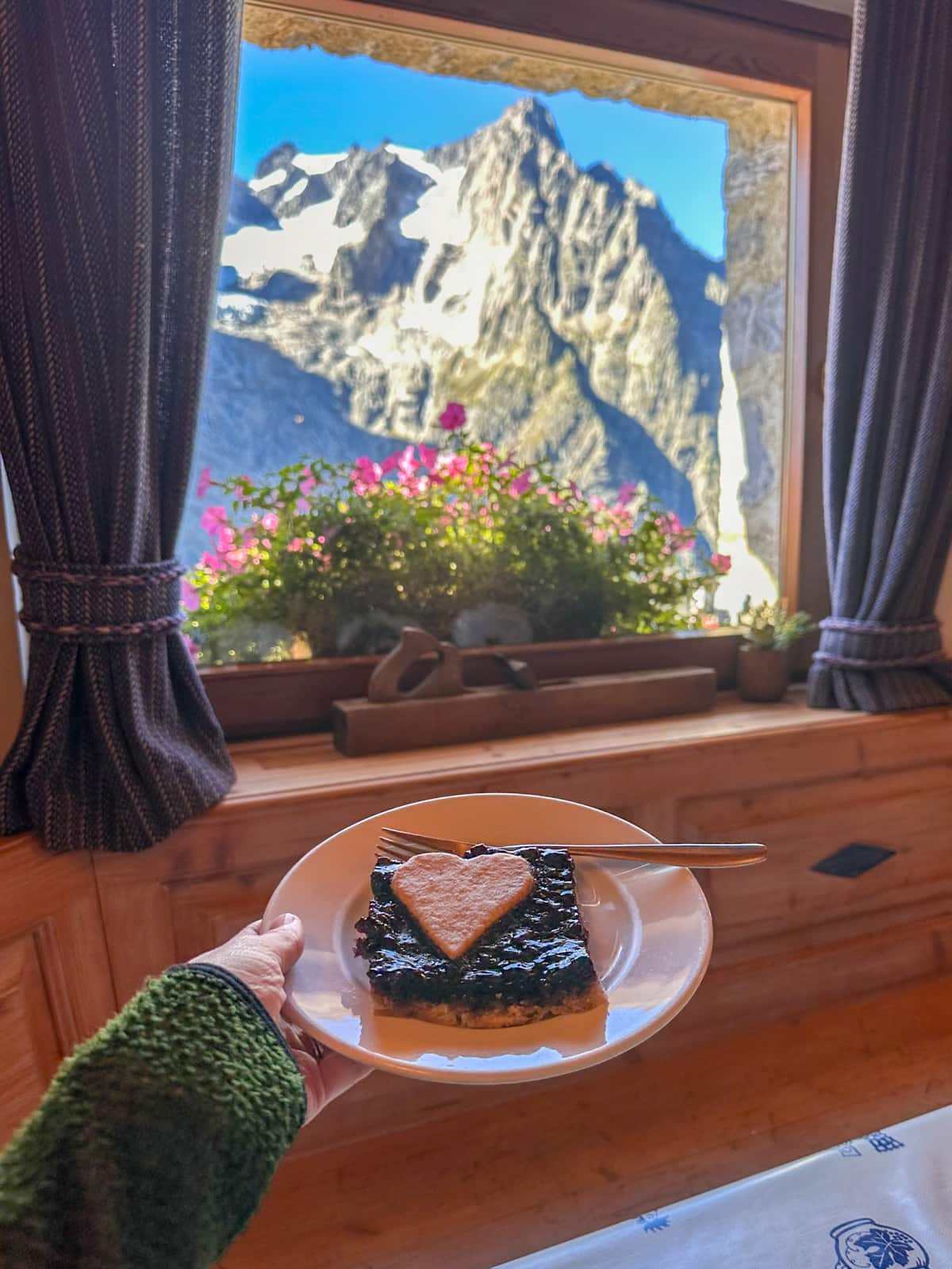 Woman holding a pice of pie in front of a window with mountains in the background in Rifugio Bonatti, Italy along the Tour du Mont Blanc