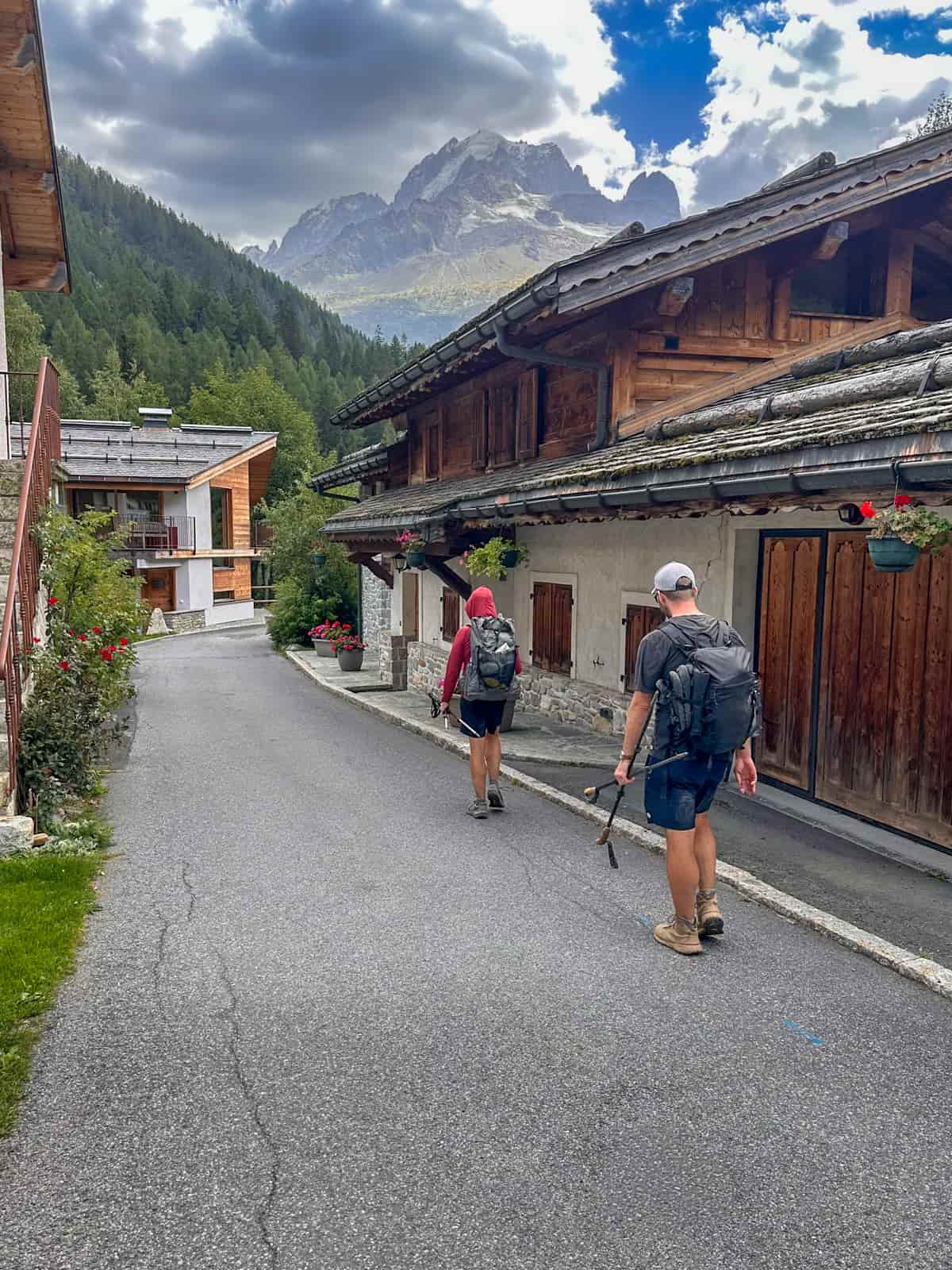 Two hikers hiking past a wooden and stone chalet in Montroc, France with the French Alps in the background of the Tour du Mont Blanc