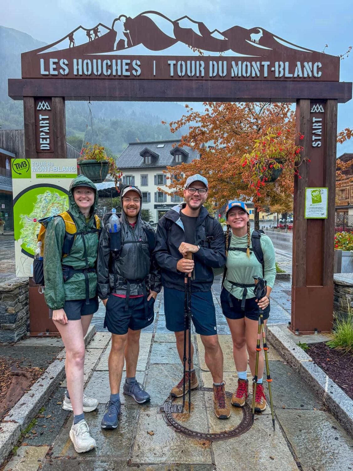 Four smiling friends standing at the Tour du Mont Blanc starting arch in Les Houches, France
