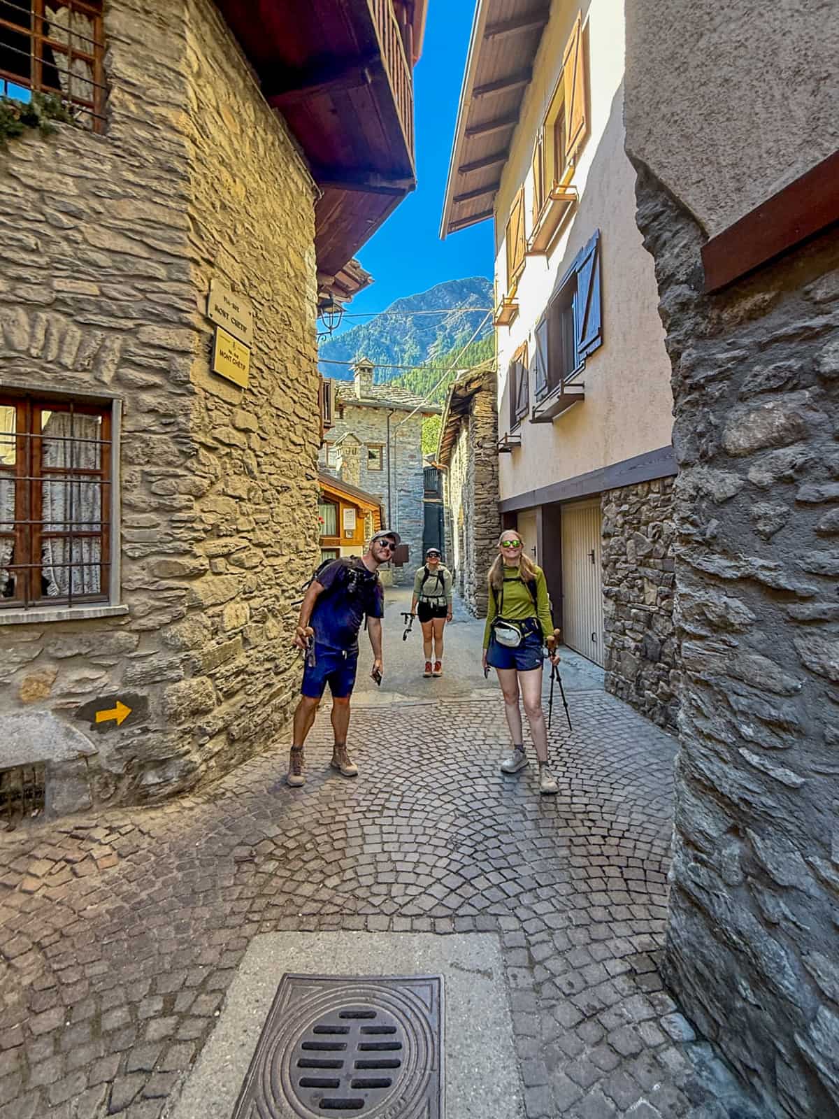 Three smiling friends standing in an alleyway between stone chalets in Courmayeur, Italy along the Tour du Mont Blanc