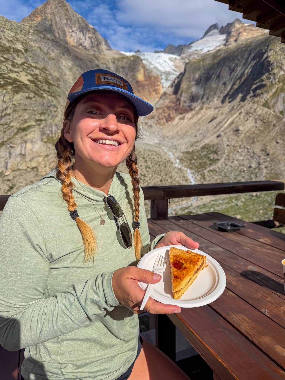 Smiling woman sitting at a picnic table and holding a piece of pie with a mountain and glacier in the background at Rifugio Elena along the Tour du Mont Blanc