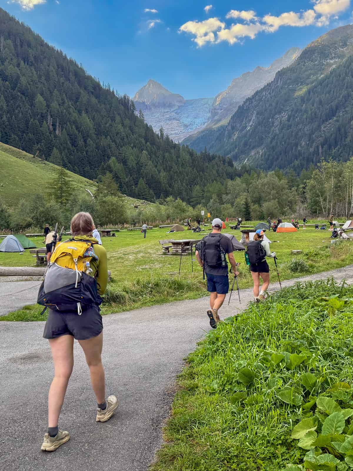 Three friends hiking along a pathway with the Le Peuty campground and Swiss Alps in the background along the Tour du Mont Blanc