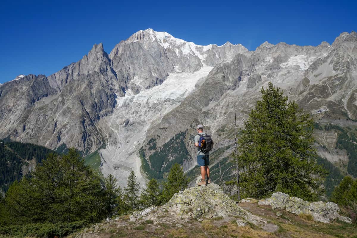 Man standing on a rocky outcropping with a glacier and Italian Alps in the background along the Val Ferret along the Tour du Mont Blanc