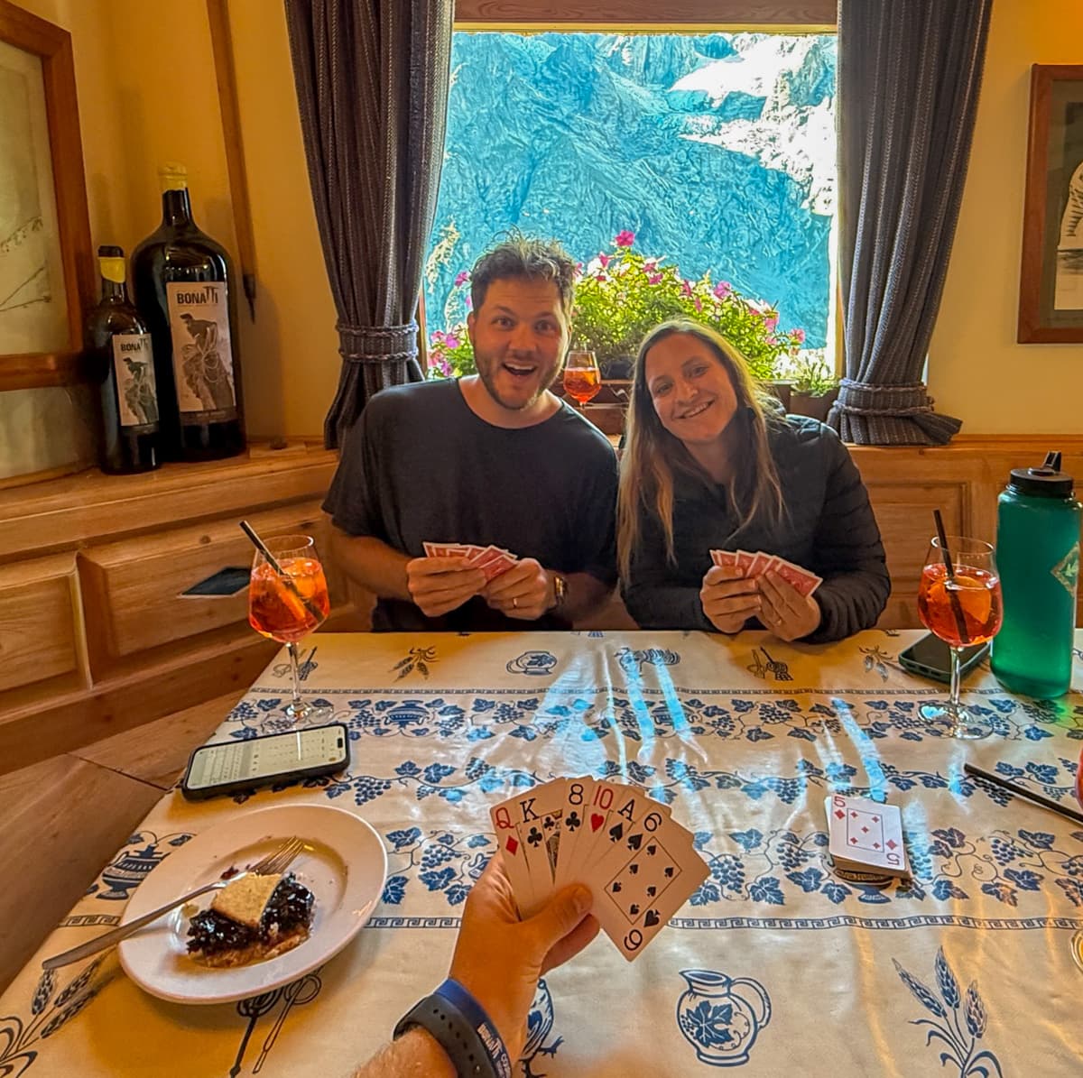 Smiling couple playing cards while eating pie and drinking an Aperol spritz at Rifugio Bonatti along the Tour du Mont Blanc