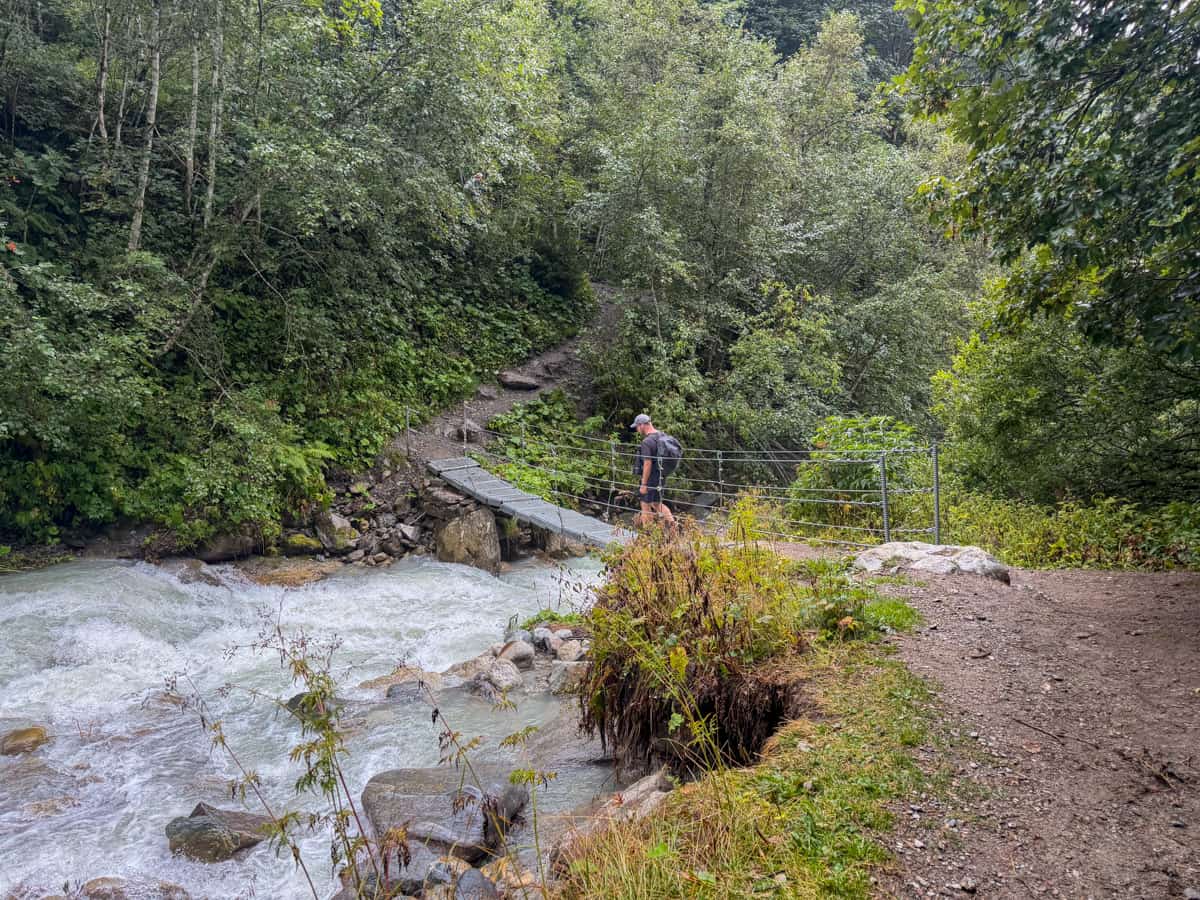 Man walking across a bridge over a rushing stream in a forest along the French portion of the Tour du Mont Blanc