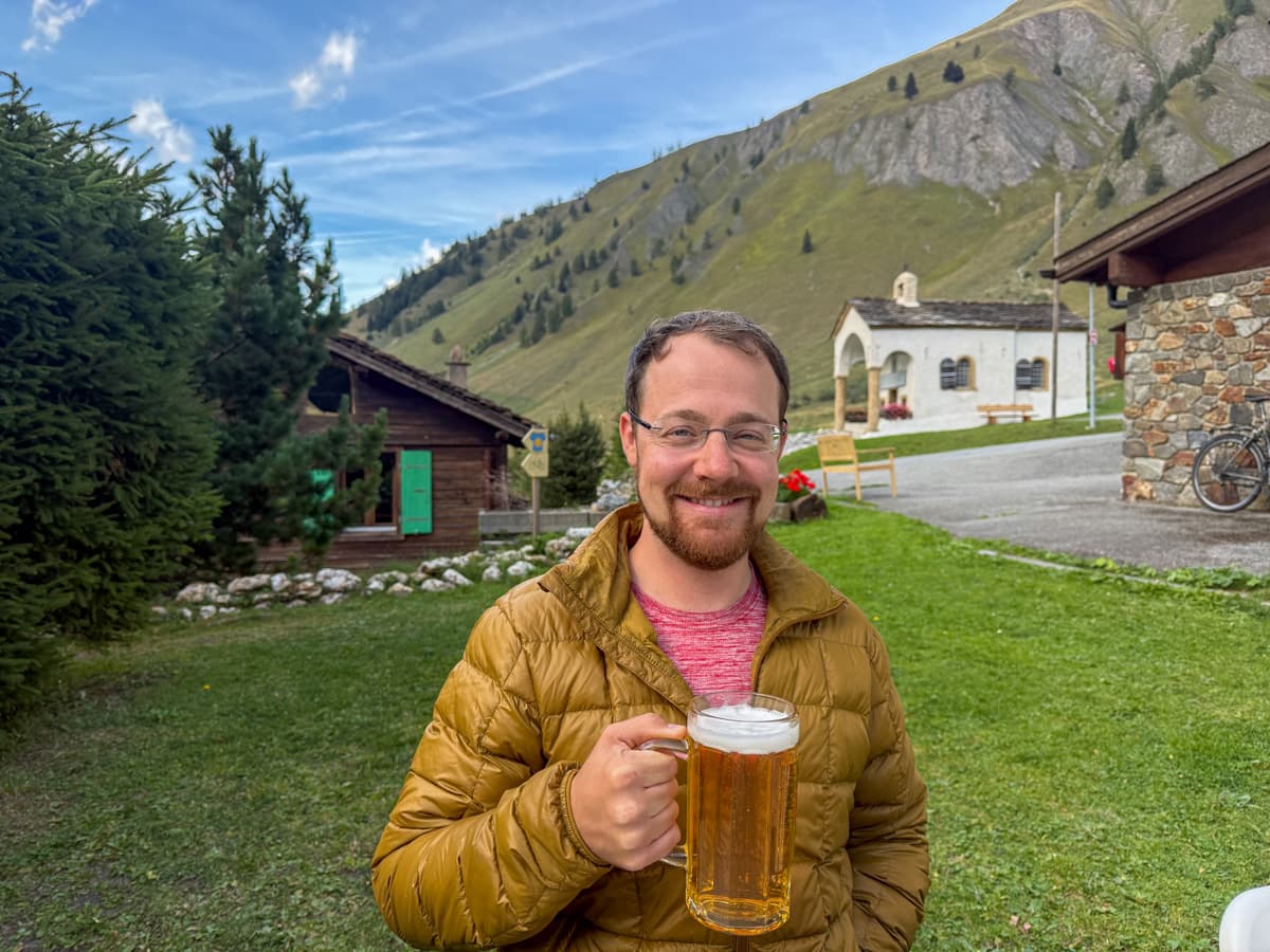 Man holding a beer with Swiss chalets in the background at Hotel du Col de Fenêtre outside of La Fouly, Switzerland along the Tour du Mont Blanc