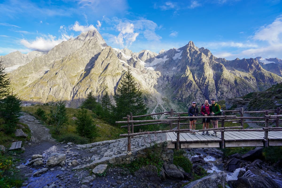 Three hikers standing on a wooden bridge with the Italian Alps in the background along the Tour du Mont Blanc