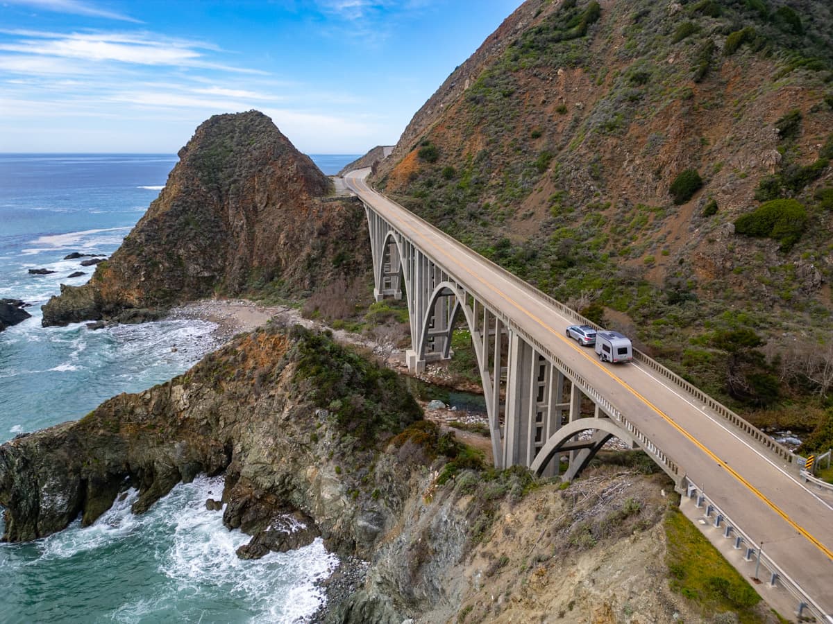 Toyota Highlander Hybrid pulling a Safari Condo Alto A1752 along California's Highway One, with rugged rock formations and waves in the background