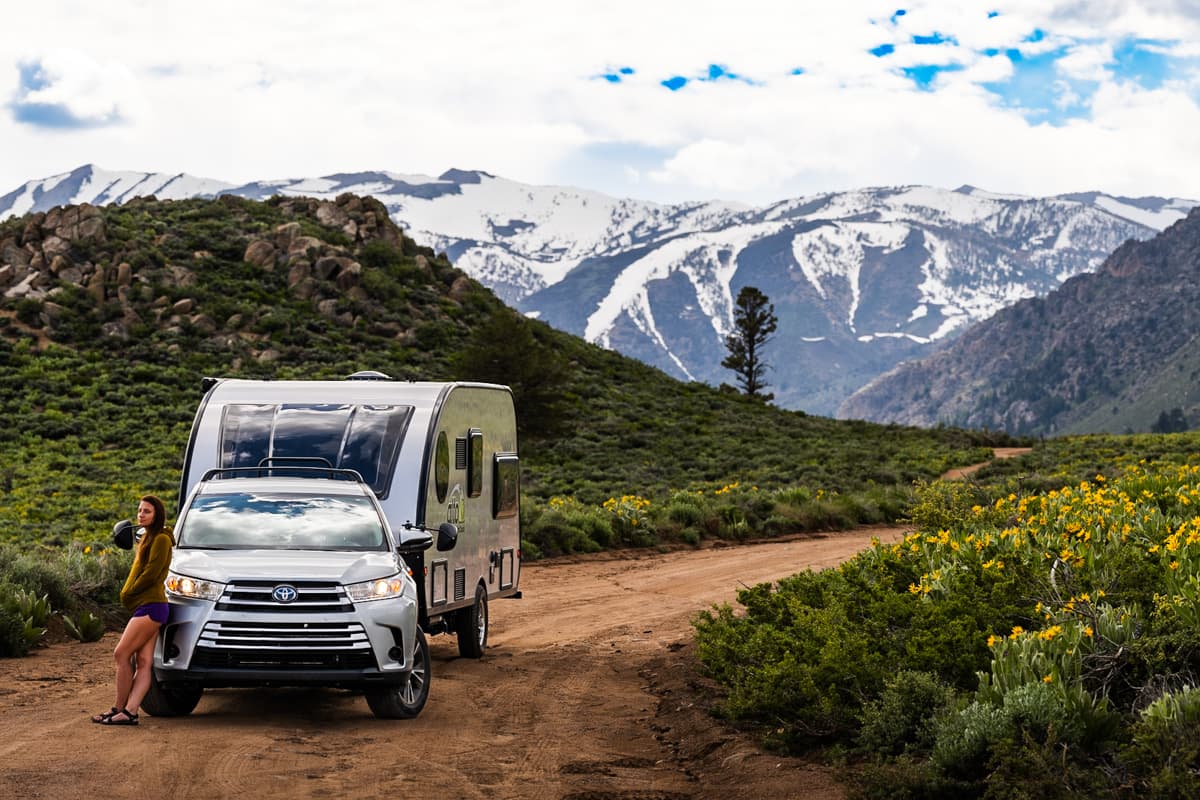 Woman leaning against a Highlander Hybrid SUV pulling a Safari Condo Alto F2114 with snowcapped mountains in the background near Bridgeport, California