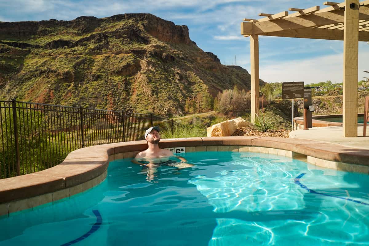 Man relaxing in a hot spring pool with red rock cliffs in the background in Zion Canyon Hot Springs in La Verkin, Utah